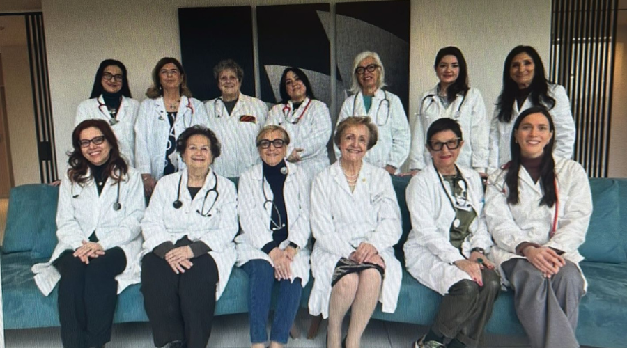 immagine di alcune donne medico (sei sedute su un divano azzurro e sette in piedi dietro di loro) che recitano nello spettacolo teatrale Le Medichesse