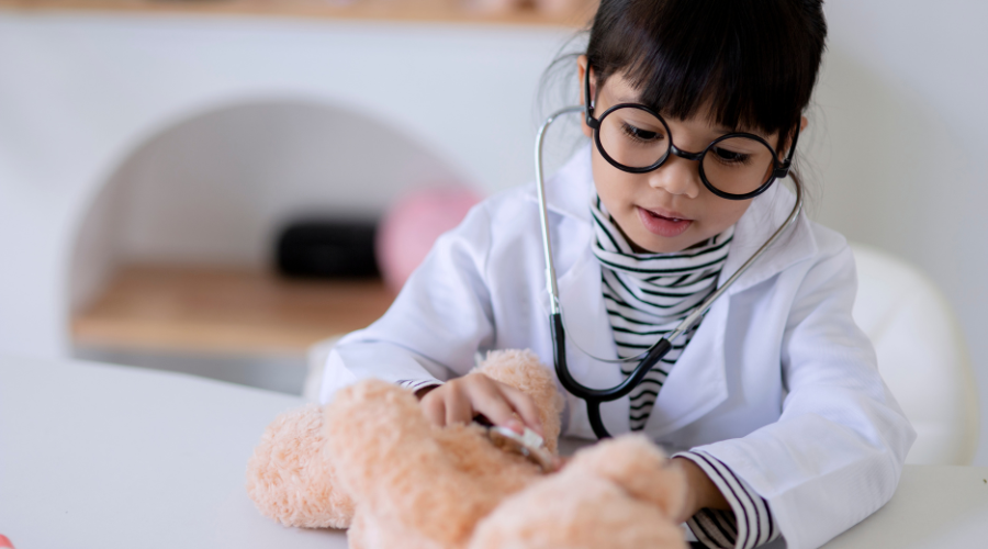 immagine di bambina in camice bianco che visita con lo stetoscopio un orsetto di peluche
