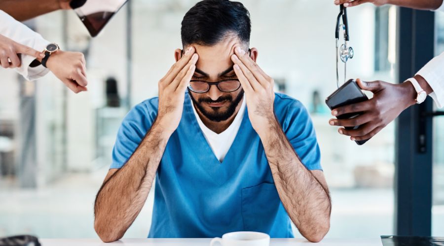 medico stanco seduto ad un tavolo sollecitato da colleghi che gli mostrano l'ora, il computer, il cellulare, lo stetoscopio, impedendogli di riposare