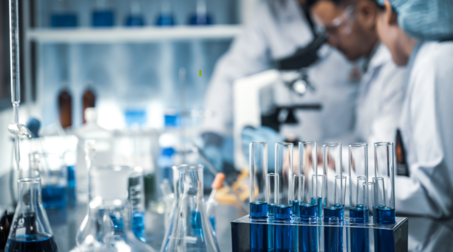 in primo piano strumenti di un laboratorio per la ricerca clinica e sullo sfondo tre operatori in camice bianco vicini ad un microscopio