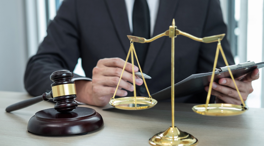 immagine di un uomo a mezzo busto in giacca e cravatta, seduto ad una scrivania. L'uomo tiene in mano una biro e dei fogli per prendere appunti. Sulla scrivania ci sono un martelletto e una bilancia simbolo della giustizia