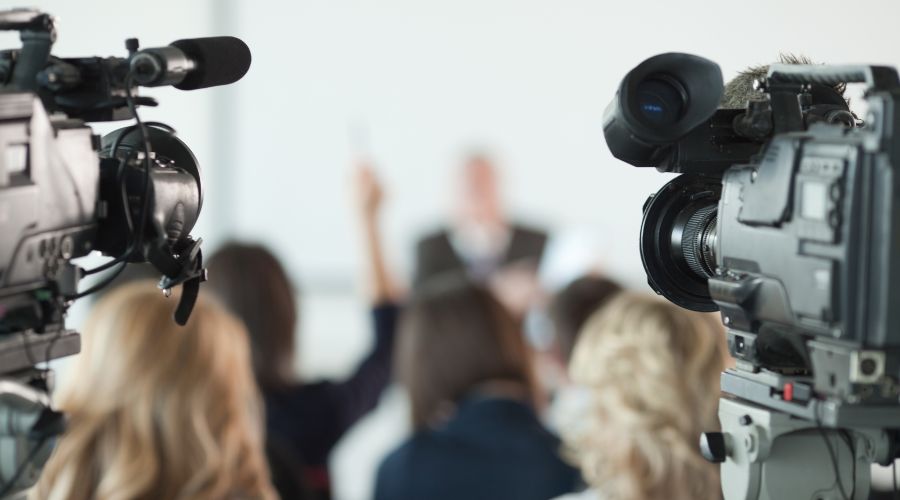 immagine di un gruppo di giornalisti (e un paio di cineprese) di spalle che partecipano ad una conferenza stampa, Uno di loro ha una mano alzata in attesa di formulare la domanda.Il gruppo ha di fronte il relatore 